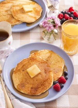 Two buttermilk pancakes on a plate with butter, syrup, and berries. More pancakes, a glass of orange juice, a small vase of flowers, and a bowl of berries in the background.