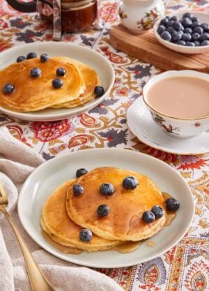 Two plates with pancakes on them, with maple syrup and blueberries on top. A cup of tea is on the side, and more berries in a bowl in the background.
