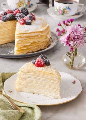 A slice of crepe cake decorated with powdered sugar and berries on a plate. The rest of the unsliced cake, cups of coffee, and a vase of flowers are in the background.