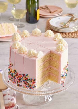 Cake on a cake stand with cuts in it demonstrating how to cut a round cake neatly and easily. Drinks in glasses and serving plates in the background.