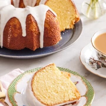 A slice of bundt cake with lemon glaze on a small plate. A cup of tea and the rest of the cake is in the background.
