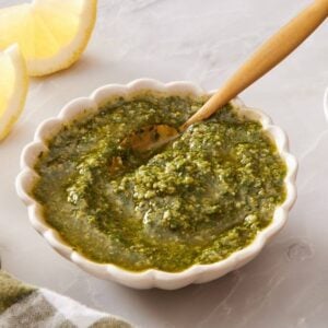 Green basil and parmesan cheese sauce in a white bowl with a spoon, surrounded by lemon wedges, salt, and black pepper.