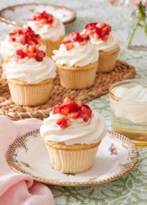 Cupcake on a plate topped with whipped topping and diced strawberries. A drink sits alongside it, and there's more strawberry shortcake cupcakes in the background.
