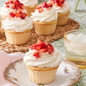 Cupcake on a plate topped with whipped topping and diced strawberries. A drink sits alongside it, and there's more strawberry shortcake cupcakes in the background.
