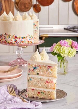 A slice of three-layer funfetti cake on a small plate with a vase of flowers right behind. The rest of the cake in the background.