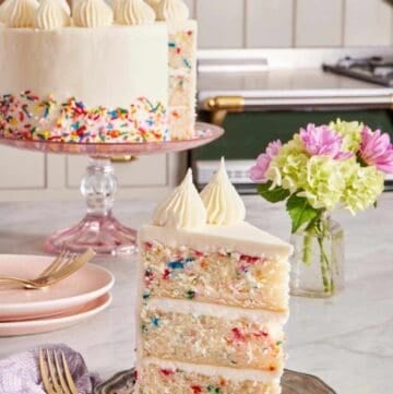 A slice of three-layer funfetti cake on a small plate with a vase of flowers right behind. The rest of the cake in the background.