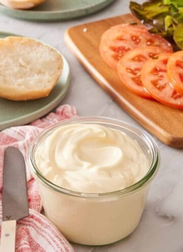 A small glass jar of mayo with sliced tomatoes and greens in the background.
