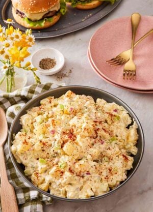 A large bowl of creamy salad with plates and burgers in the background.
