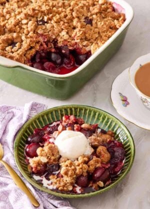 Cherry crisp in a green bowl with a scoop of vanilla ice cream on top. A cup of tea and a baking dish containing the rest of the crisp is in the background.