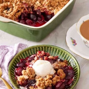 Cherry crisp in a green bowl with a scoop of vanilla ice cream on top. A cup of tea and a baking dish containing the rest of the crisp is in the background.