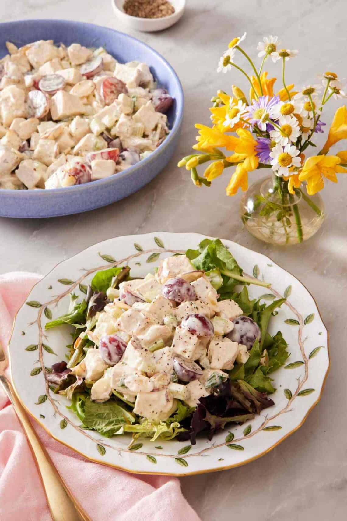 Chicken salad on mixed greens on a patterned plate with a vase of flowers and more salad alongside.
