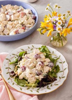Chicken salad on mixed greens on a patterned plate with a vase of flowers and more salad alongside.