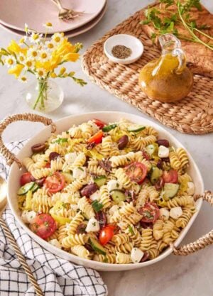A deep dish of pasta-based salad with a small vase of flowers alongside. A small jar of salad dressing is next to it.