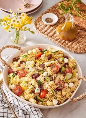 A deep dish of pasta-based salad with a small vase of flowers alongside. A small jar of salad dressing is next to it.