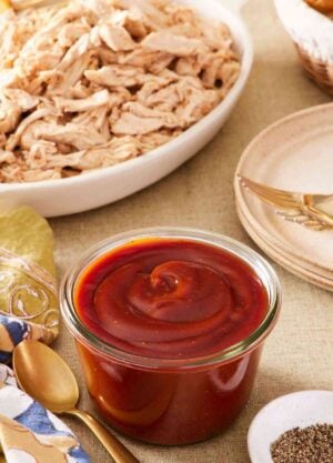 Homemade barbecue sauce recipe in a small glass container with a spoon next to it. Shredded meat, black pepper, and plates around it.