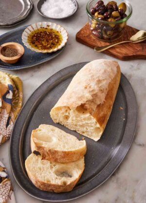 A loaf of ciabatta bread with two slices cut out, with olives and olive oil in the background.