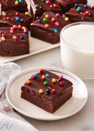 Cosmic brownie on a white plate next to a side of milk, with a platter of them in the background.
