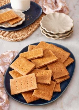 Graham crackers on a blue platter with small serving plates, more crackers, and a glass of milk in the background.