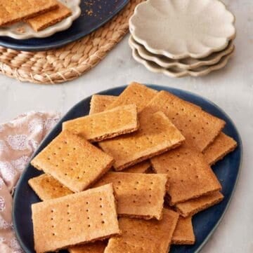 Graham crackers on a blue platter with small serving plates, more crackers, and a glass of milk in the background.