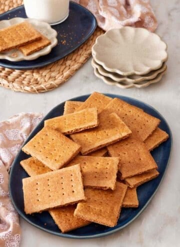 Graham crackers on a blue platter with small serving plates, more crackers, and a glass of milk in the background.