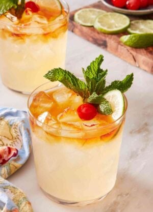 Easy mai tai recipe garnished with lime, mint, and cherry. Another cocktail and a wooden board with cocktail garnishes in the background.