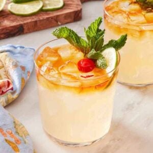 Two mai tais on a marble countertop with a table linen on the side, as well as a small dish of cherries. Wooden board with lime wheels and wedges in the background.