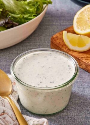 Ranch dressing in a small glass jar with sliced lemons and greens in the background.