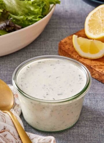 Ranch dressing in a small glass jar with sliced lemons and greens in the background.