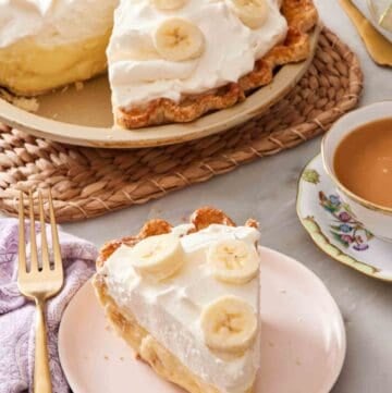 A slice of banana cream pie next to a cup of tea, with the rest of the pie in the background.