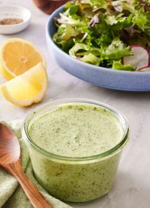 A glass jar filled with creamy green goddess dressing with sliced lemon and salad greens in the background.