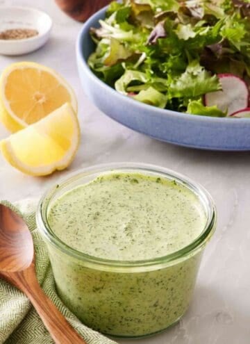 A glass jar filled with creamy green goddess dressing with sliced lemon and salad greens in the background.