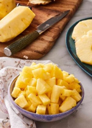 Diced pineapple in a bowl. Wooden cutting board with more pineapple in the background.