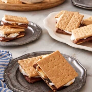 Two s'mores on a plate with more s'mores and ingredients for how to make s'mores in the background.