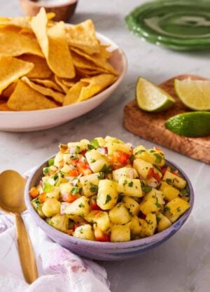 A heaped bowl of pineapple salsa on a countertop with lime wedges and chips in the background.