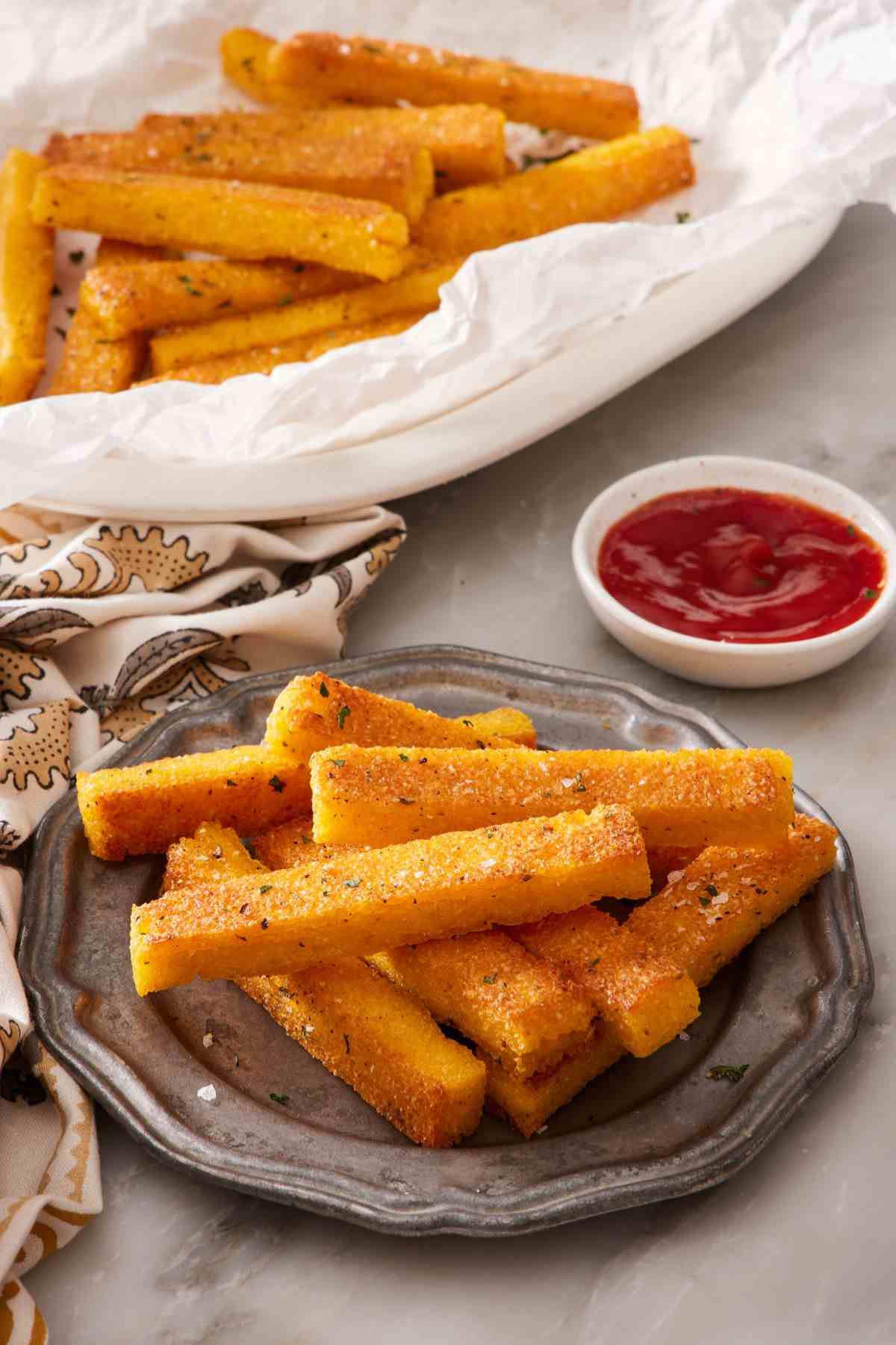 Homemade polenta fries on a small plate next to a small dish of ketchup. More fries in the background.