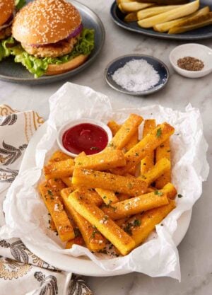 A dish of polenta fries on a paper towel with a burger in the background.