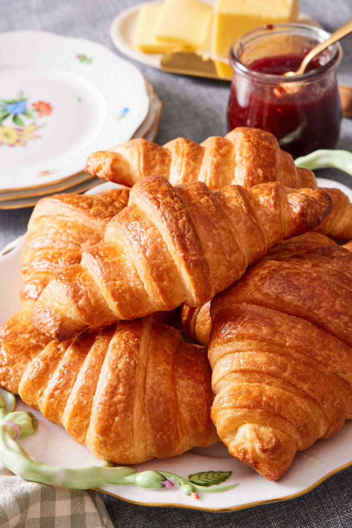 A pile of homemade croissants on a plate with jam and butter in the background.