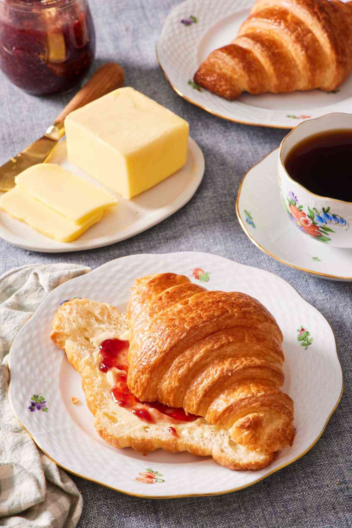 A croissant on a plate, halved and smeared with jam and butter. Coffee, butter, and another croissant also in frame.