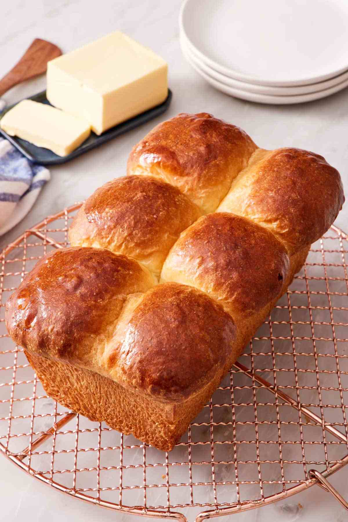 A loaf of brioche bread on a wire rack with butter in the background.