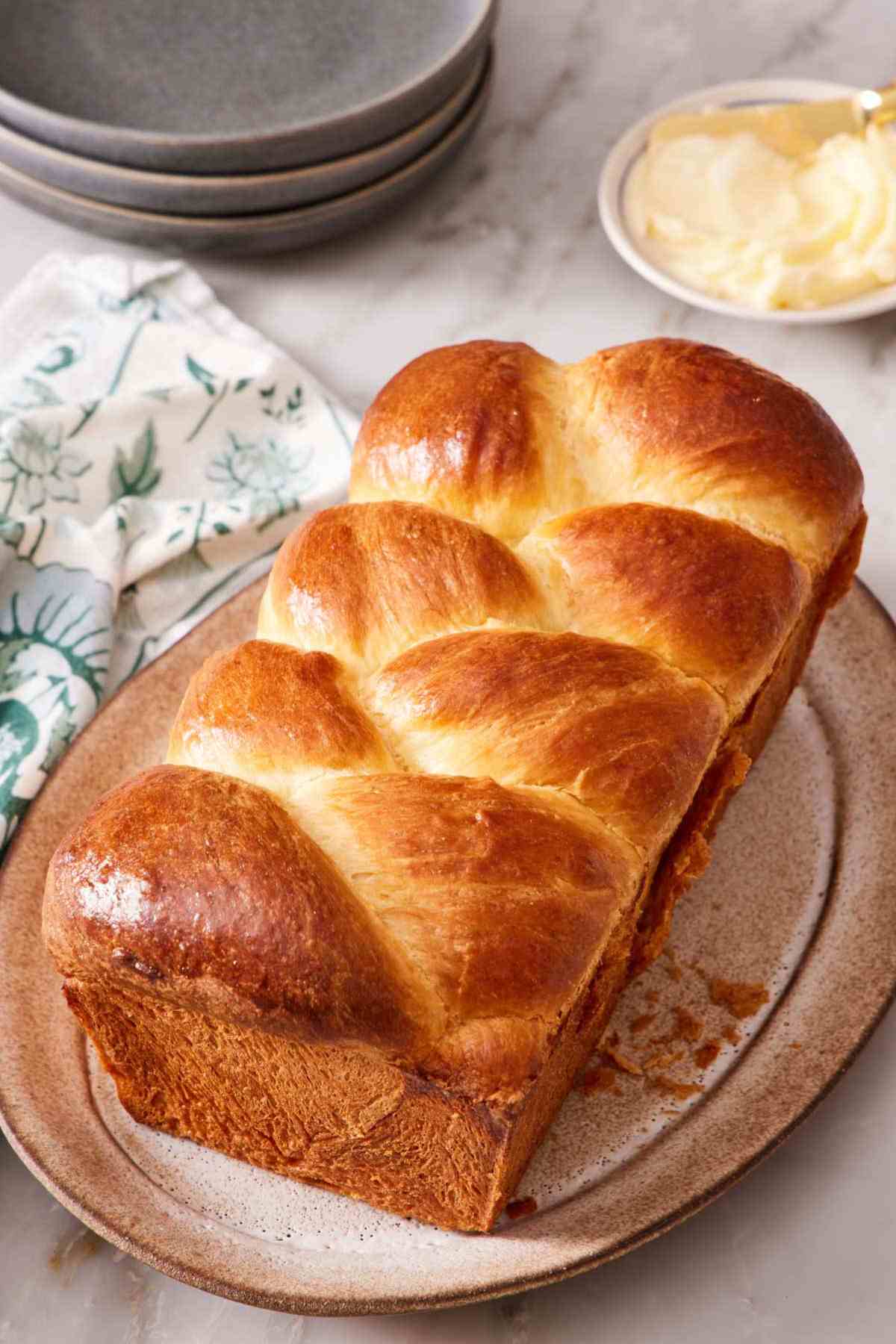 A loaf of braided brioche on a platter with butter and small plates in the background.