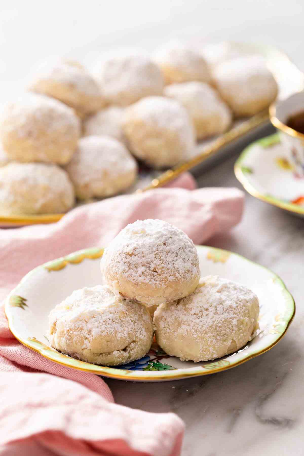 A pile of 3 Mexican wedding cookies (polvorones) on a small plate, with more cookies on a platter in the background.