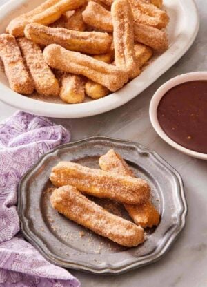 Air fryer churros on a plate, with more on a platter behind it, as well as a bowl of chocolate dipping sauce.