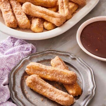 Air fryer churros on a plate, with more on a platter behind it, as well as a bowl of chocolate dipping sauce.