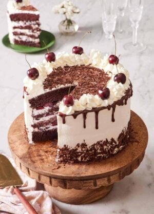 A black forest cake on a wooden cake stand with a slice cue out. The slice is on a small serving plate in the background.