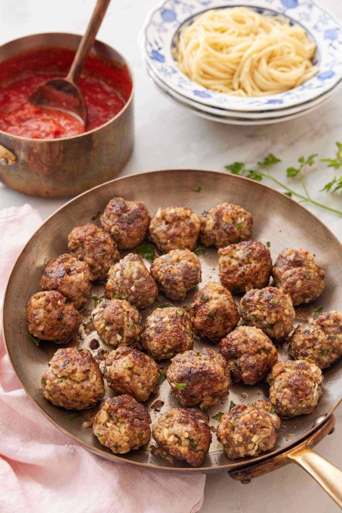 A skillet of homemade meatballs with sauce and pasta in the background.