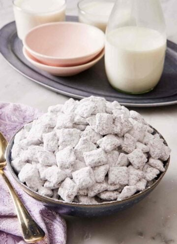 A bowl piled high with homemade puppy chow and a jug of milk with glasses and small bowls in the background.