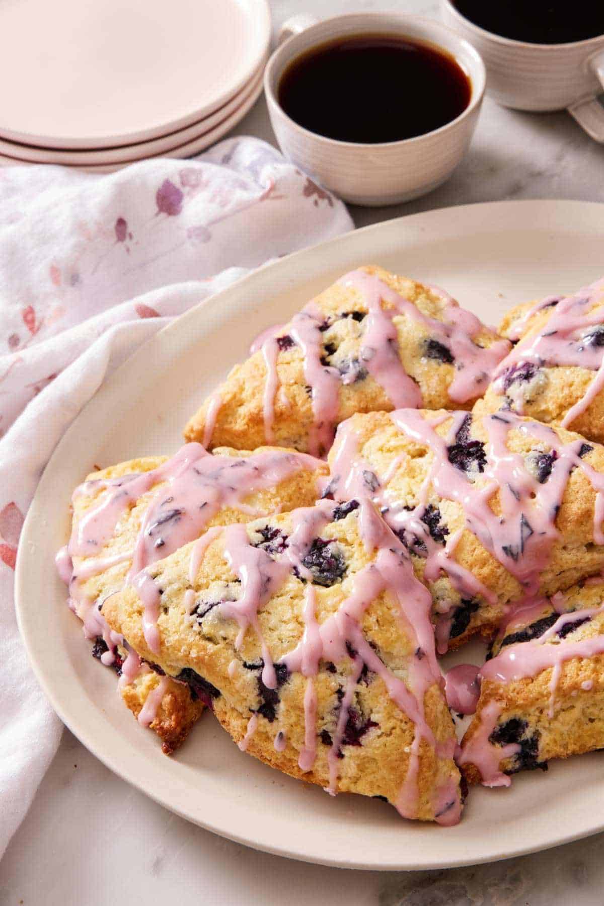 Blueberry scones topped with blueberry glaze on a platter alongside a cup of coffee.
