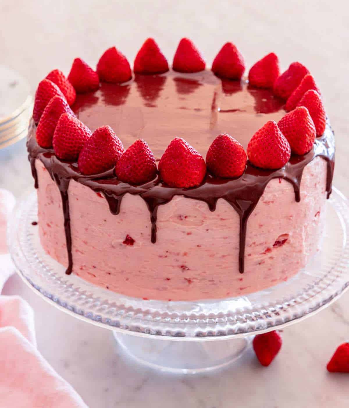 A chocolate strawberry cake topped with ganache and fresh strawberries on a cake stand.
