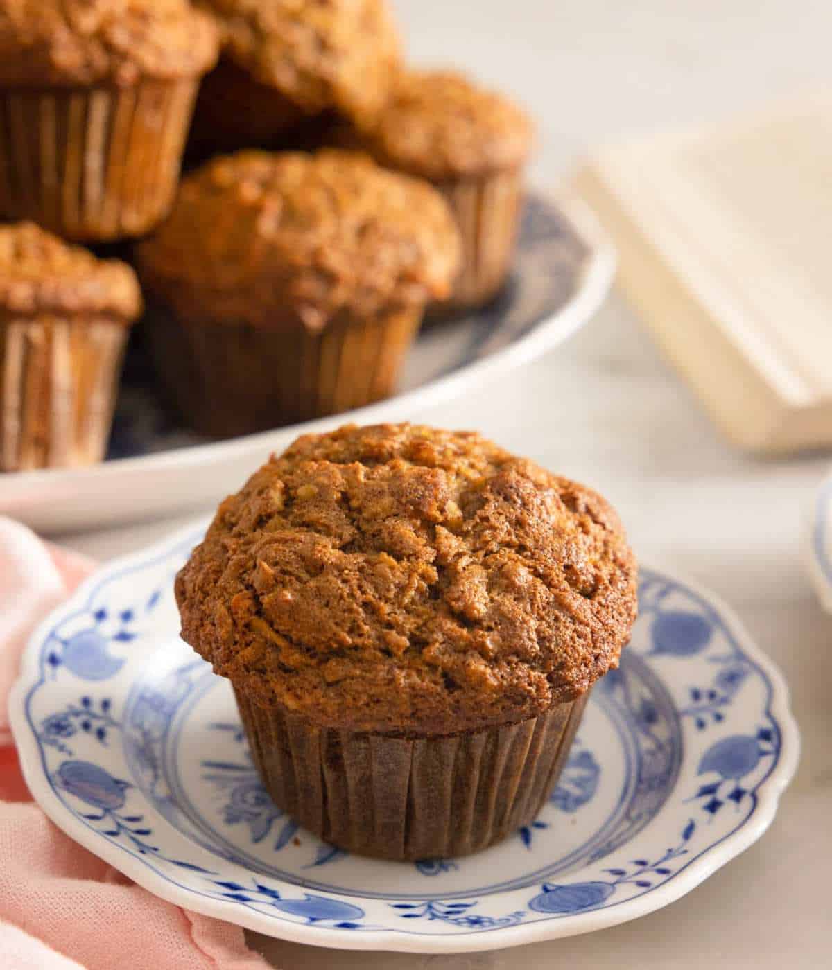 A morning glory muffin on a patterned plate with more muffins piles on a platter in the background out of focus.
