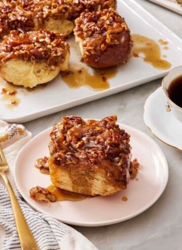 A pecan sticky bun on a white plate with the rest of the sticky buns mostly out of frame.